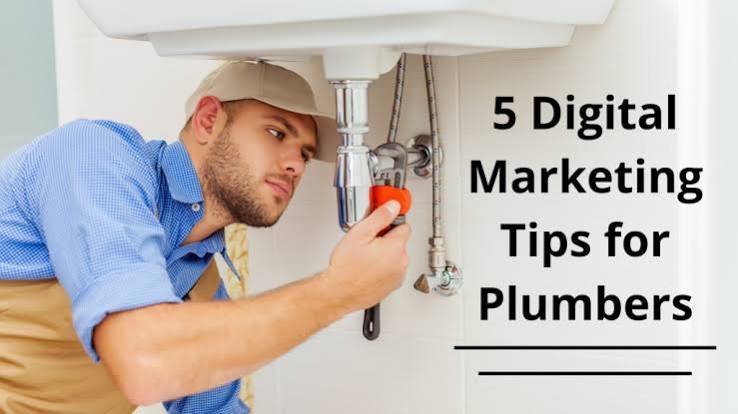 A plumber wearing a light blue work shirt, beige apron, and tan cap inspects the plumbing beneath a kitchen sink using a red-handled tool. On the right side, bold black text reads, “5 Digital Marketing Tips for Plumbers.” The image combines a real-life plumbing scene with a clean, minimalist design to illustrate an article about digital marketing strategies for plumbing businesses.